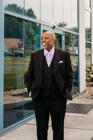 Smiling bald groom in dark suit with lavender vest posing outside glass building.