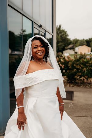 A radiant bride in an off-shoulder white gown and veil smiles outdoors on her wedding day.