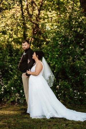 Bride in white gown with veil and groom in dark suit embrace outdoors surrounded by lush green trees.