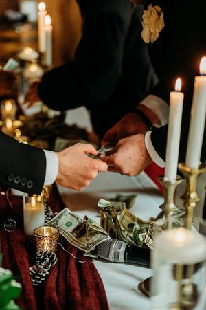 Hands exchanging money over a candlelit wedding reception table with elegant gold decor.