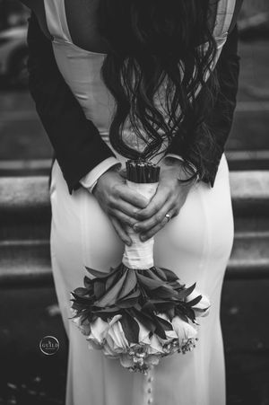 Black and white photo of bride and groom holding a white rose bouquet together from behind.