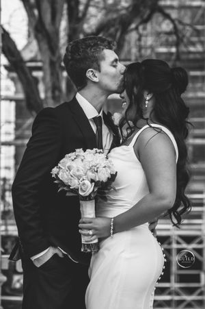Black and white photo of newlyweds sharing a kiss, bride holding white rose bouquet outdoors.