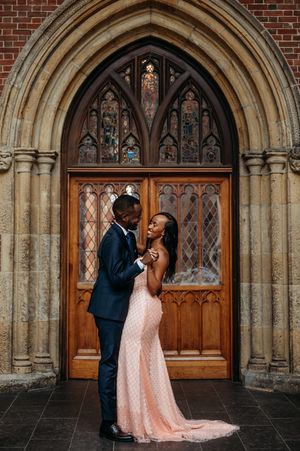 Couple in elegant wedding attire embracing before ornate Gothic church doors with stained glass windows.