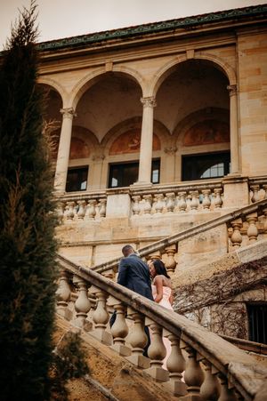 Couple ascending ornate stone staircase of a historic European villa with arched colonnade and decorative balustrade.