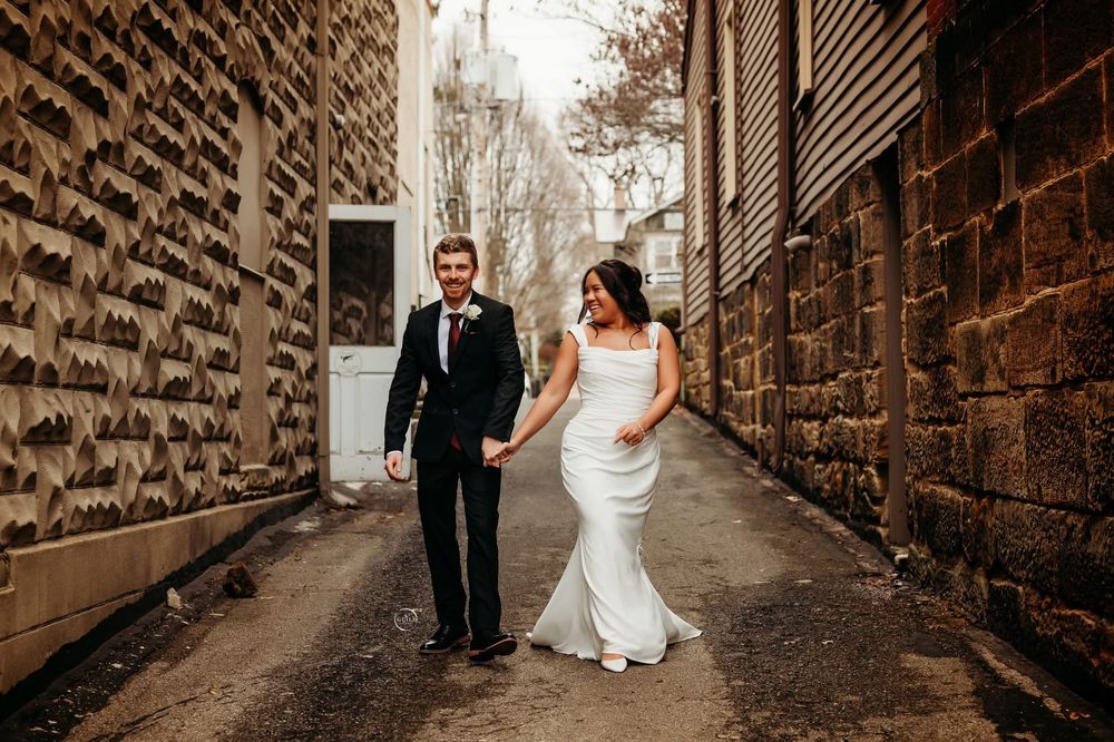 Bride in white gown and groom in black suit walk hand-in-hand down a rustic stone alleyway.