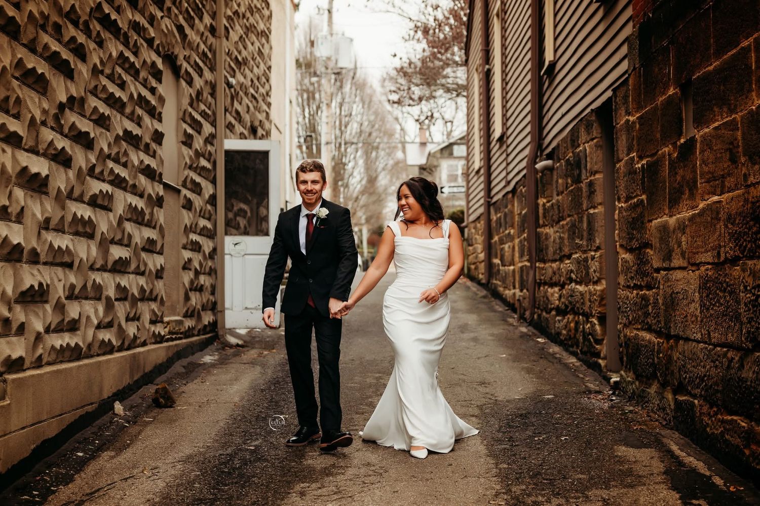 Bride in white gown and groom in black suit walk hand-in-hand down a rustic stone alleyway.