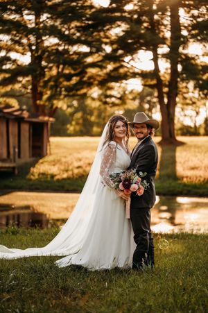 A bride and groom pose romantically at golden hour near a pond surrounded by tall trees.
