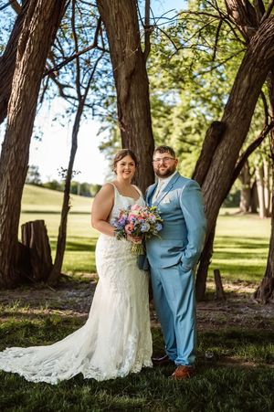 Bride in lace gown and groom in light blue suit pose with colorful bouquet among trees outdoors.