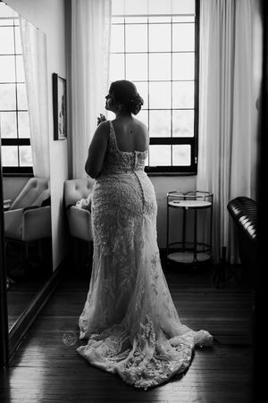 Bride in lace wedding gown with train stands by window in black and white portrait photo.