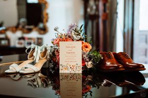 Wedding flat lay with floral bouquet, invitation, bridal heels, and brown leather Oxford shoes on a reflective surface.
