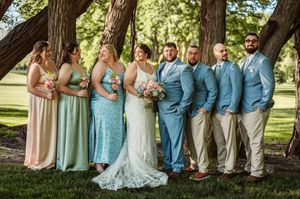 Wedding party poses under trees, bridesmaids in sage green gowns and groomsmen in blue suits flanking the bride and groom.