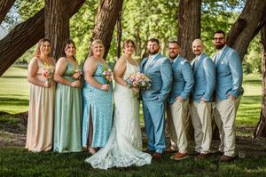 Wedding party poses outdoors under trees, bridesmaids in pastel gowns, groomsmen in blue suits flanking bride and groom.