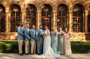 Wedding party posing in blue suits and sage bridesmaid gowns before ornate arched brick building windows.