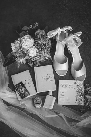 Black and white flat lay of wedding details including bouquet, heels, vow books, and invitation.