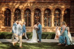 Bride and groom kissing outside historic brick building with arched windows while wedding party walks past in motion blur.