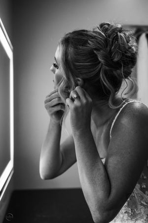A bride adjusts her earring while gazing into a lit vanity mirror in black and white.