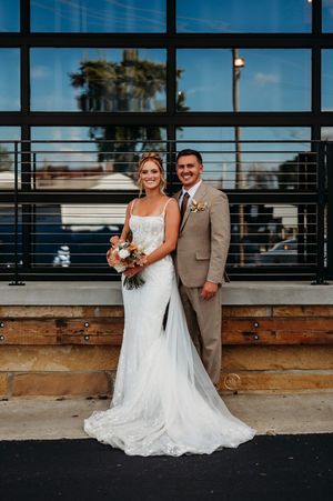 A smiling bride in a fitted lace gown and groom in a tan suit pose before large windows.