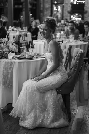 A bride in a lace gown sits smiling at the sweetheart table during her wedding reception.