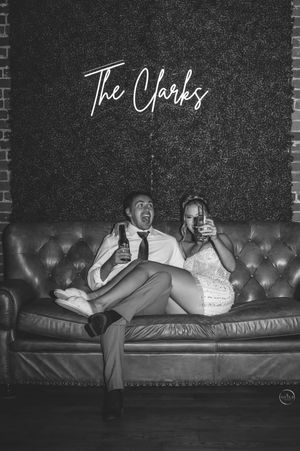 Newlyweds relax on a leather sofa beneath a neon sign reading The Clarks at their reception.