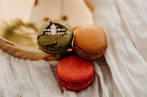 Three colorful macarons in green, orange, and red displayed with a diamond engagement ring on top.