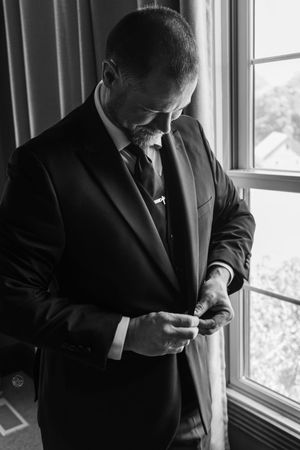 Black and white photo of a groom in a suit adjusting his cufflinks by a window before his wedding.