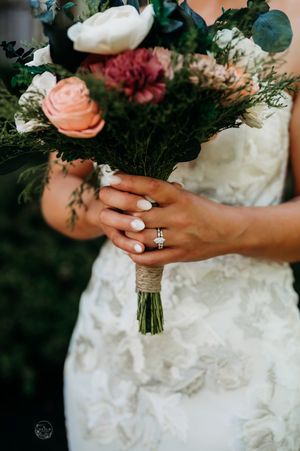Bride in lace gown holding bouquet of pink, white, and mauve flowers with greenery, showcasing diamond engagement ring.