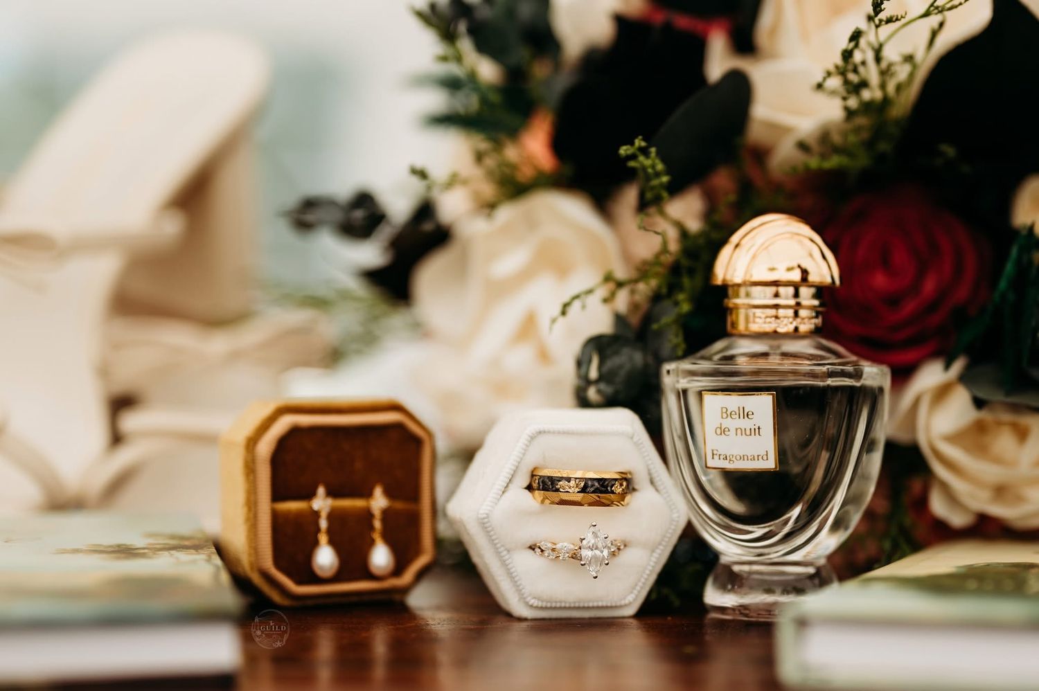 Elegant wedding flat lay with gold earrings, diamond ring, perfume bottle, and floral bouquet on a wooden surface.