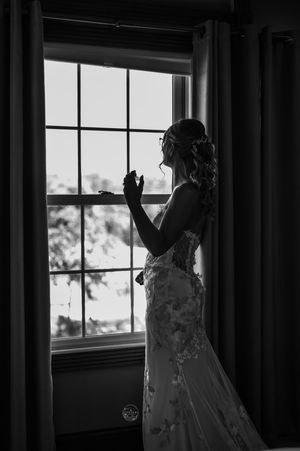 Bride in lace wedding gown stands silhouetted by window light in elegant black and white bridal portrait.