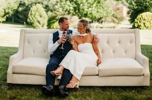 Bride and groom sitting on elegant white tufted sofa on grass, toasting with wine glasses at outdoor wedding reception.
