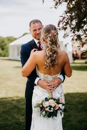 Bride and groom embrace outdoors, she holds a rose bouquet in a lace gown as he smiles in a navy suit.