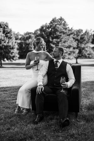 Black and white wedding photo of bride and groom sitting in leather chair outdoors, holding champagne glasses.