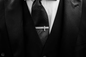 Black and white close-up of a man in a tuxedo wearing a patterned tie with a silver cross tie clip.