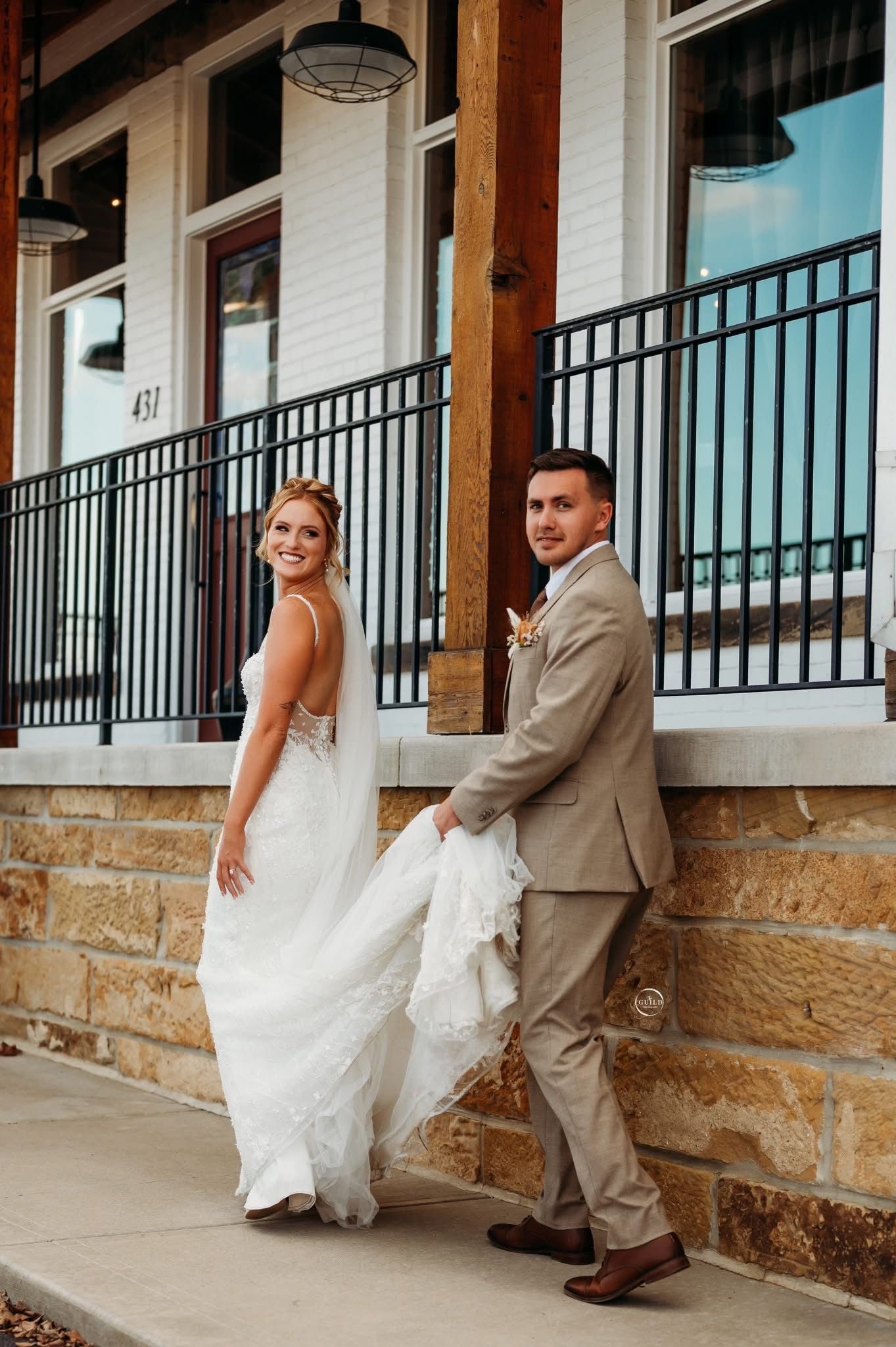 A bride lifts her gown train while smiling back at the camera as the groom leans on stone wall.