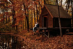 Couple sitting on dock beside rustic cabin surrounded by vibrant autumn foliage reflected in pond.