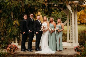 Wedding party poses under pergola in sage bridesmaid dresses and black suits amid autumn blooms.