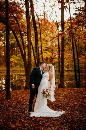 Bride and groom share romantic kiss in forest surrounded by golden fall leaves near pond.