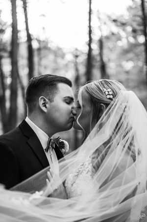 Black and white portrait of bride and groom kissing under flowing veil in forest setting.