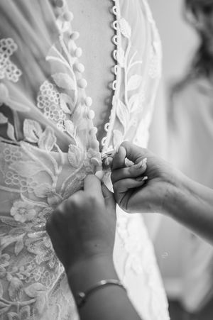 Black and white close-up of hands fastening delicate lace buttons on elegant bridal gown.