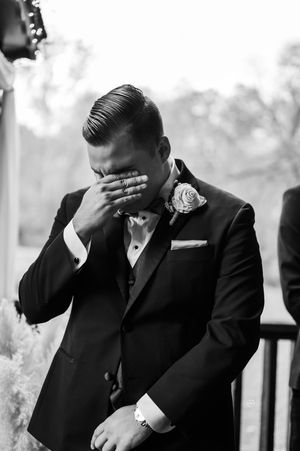 Emotional groom in black tuxedo covering face with hand during touching wedding ceremony moment.