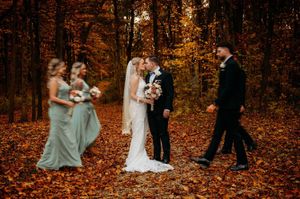 Bride and groom kiss while wedding party walks through stunning autumn forest of golden leaves.