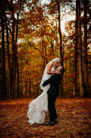 Groom dramatically dips bride in white gown for kiss among vibrant fall foliage in forest.