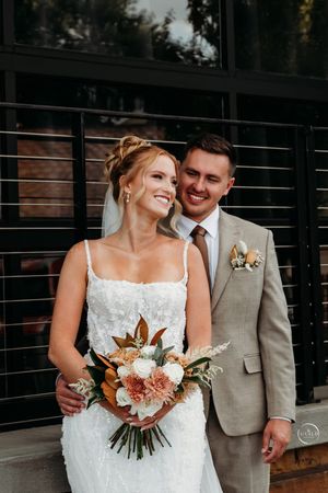 Smiling bride in white gown holds warm-toned bouquet beside groom in tan suit outdoors.