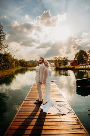 Bride and groom stand back-to-back on wooden dock at sunset with serene lake reflection.