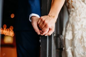 Close-up of bride and groom holding hands, bride wearing a delicate gold bracelet on their wedding day.
