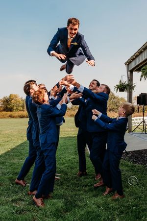 Groomsmen tossing the groom into the air in matching navy suits at an outdoor wedding celebration.
