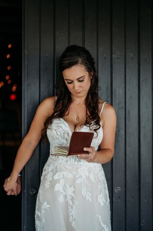 Beautiful bride in lace wedding gown reading vows from a small brown leather book against dark wall.