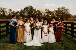 Bride posing with bridesmaids in colorful mismatched gowns by a wooden fence at fall wedding.