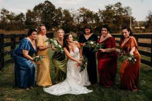 Bride laughing with bridesmaids in jewel-toned velvet gowns holding bouquets at outdoor fall wedding.