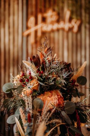 Lush boho wedding floral arrangement with burgundy blooms and pampas grass beneath a glowing neon sign.