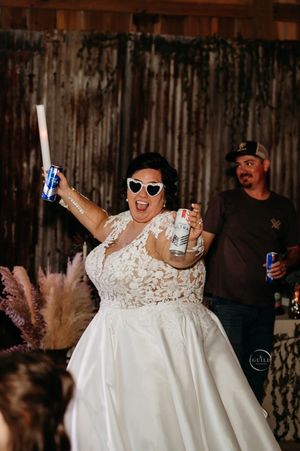 Joyful bride in lace gown and heart sunglasses celebrates at rustic barn wedding reception with drinks.
